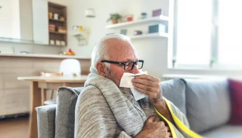 Älterer Mann zu Hause auf dem Sofa und in eine Decke eingewickelt, der hustet und sich dabei ein Taschentuch vor den Mund hält.