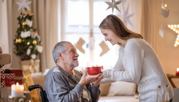 Junge Frau, die dem älteren Großvater im Rollstuhl zu Hause zu Weihnachten eine Tasse Tee gibt.