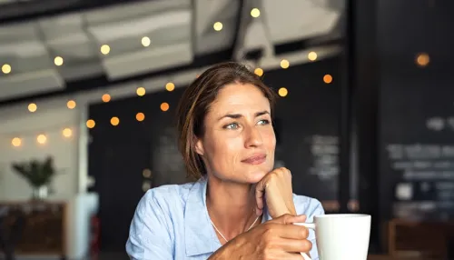 Ältere Frau sitzt im Café, blickt nachdenklich und hält einen Kaffeebecher in der Hand.