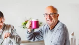 Ein älterer Mann mit weißem Haar, der ein blau kariertes Hemd trägt, sitzt auf einem Sofa und schaut nach rechts. Ihm gegenüber steht eine Frau, die ein graues Hemd trägt. Sie reicht ihm einen weißen Teller, auf dem ein in zwei Hälften geschnittener, gelber Apfel liegt.