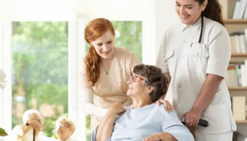 Zwei Pflegerinnen kümmern sich lächelnd um eine ältere Frau im Rollstuhl. Die Szene spielt in einem hellen, freundlichen Raum mit großen Fenstern im Hintergrund. Die Atmosphäre wirkt warm und fürsorglich, was auf eine angenehme Pflegeumgebung hinweist.